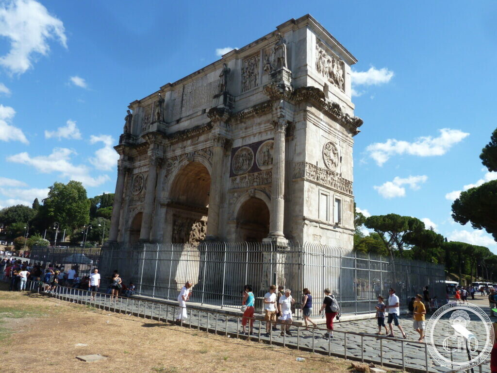 The Arch of Constantine seen at an angle, its side and front both visible, a railing and visitors in front, trees of the Palatine behind