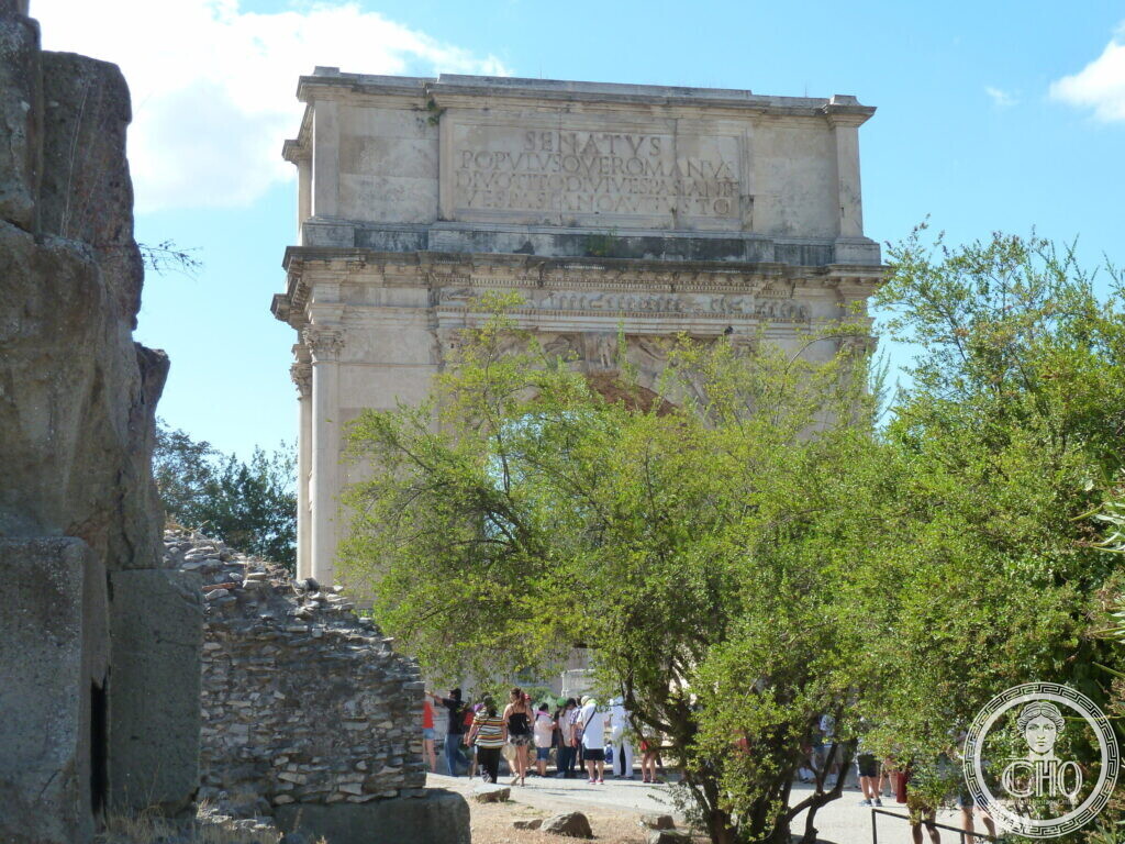 The upper part of a single marble arch with a Latin dedication to the deified Titus on the attic, framed by a tree and a ruined brick wall