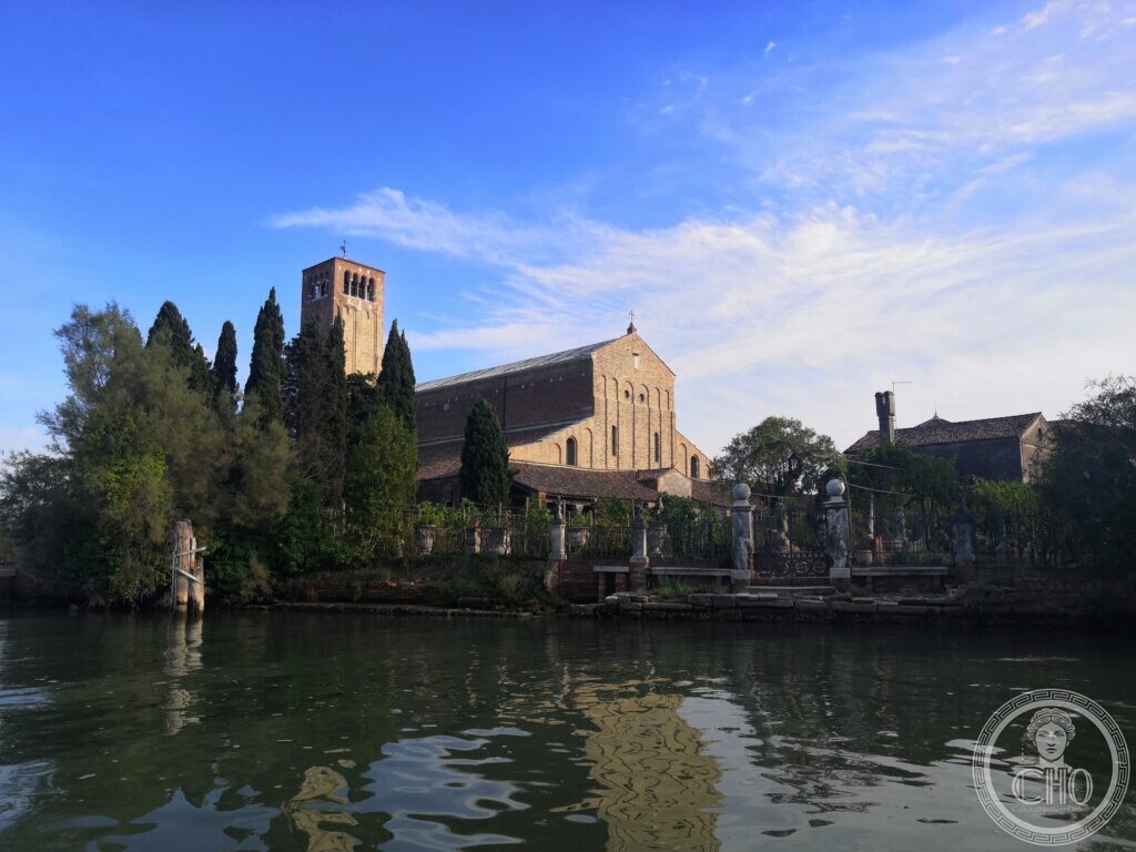 A tall brick basilica with a plain gabled front and a square bell tower with paired openings, seen across a canal, with cypresses, a vineyard behind an iron fence and a low house to the right