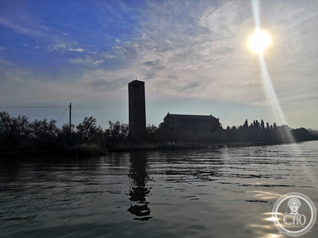 The silhouette of a tall square bell tower and a long church roof against a bright sky with the sun above, reflected in the dark water of a canal