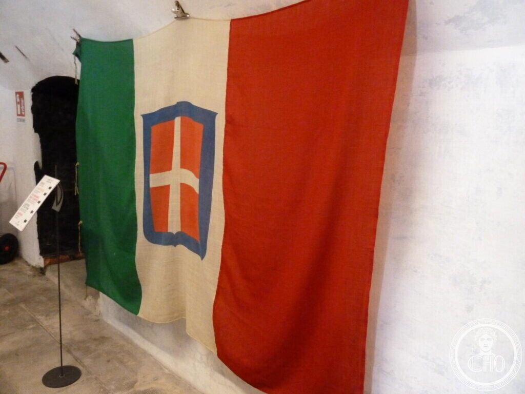 A large green white and red flag with a crowned shield bearing a white cross, hanging on the wall of a whitewashed vaulted room