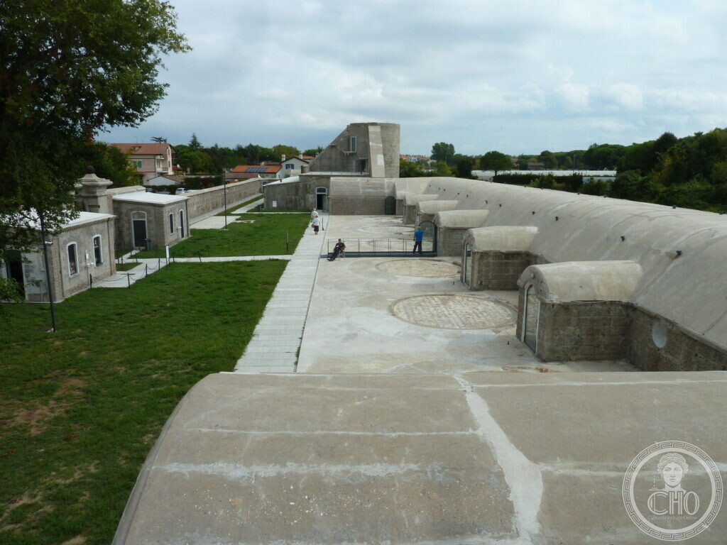 A long low concrete fortification seen from above, with barrel-vaulted roofs, circular gun platforms set into the paving and a lawn with small stone outbuildings alongside