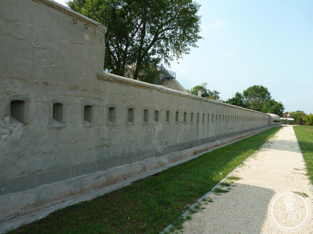 A long concrete wall curving away into the distance with a regular row of small rectangular loopholes, a gravel path beside it