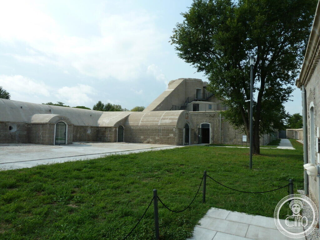 A row of earth-covered barrel-vaulted concrete casemates with arched doorways, a lawn and a tree in front, a square turret rising behind