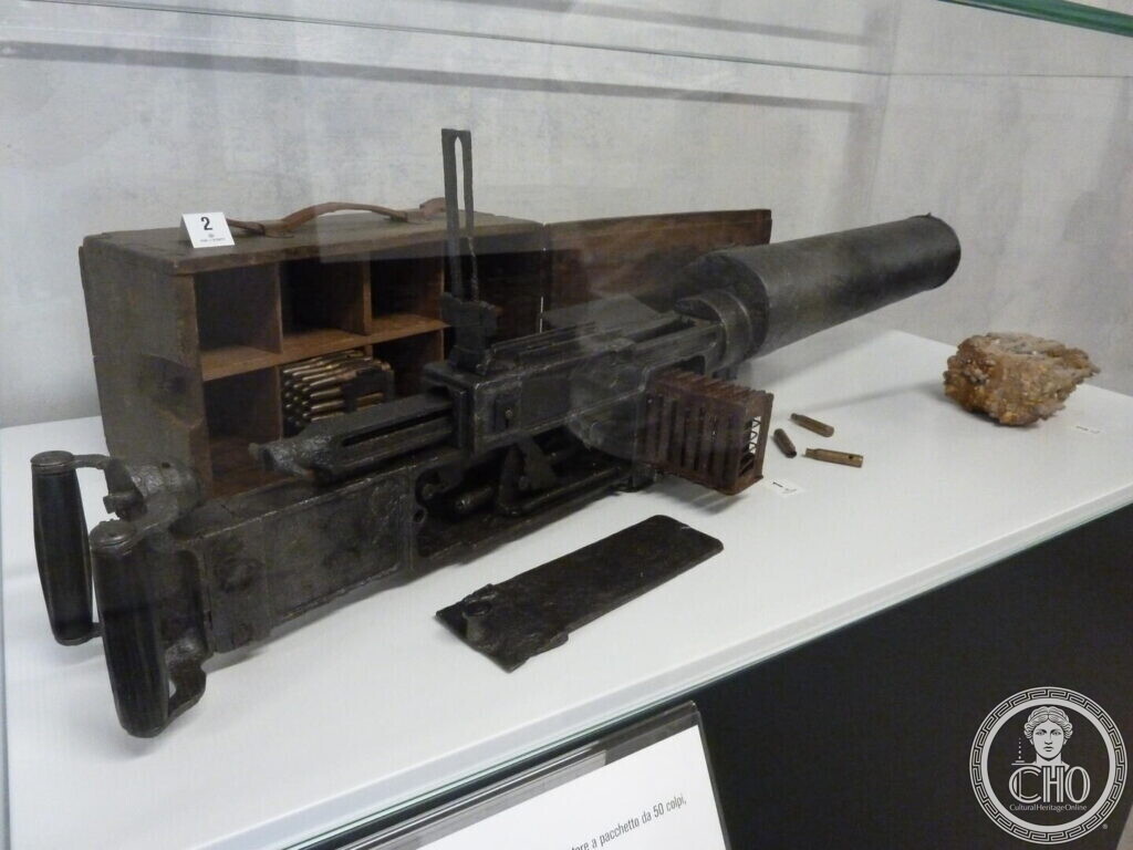 A heavy machine gun with a wooden ammunition box, cartridge strips and spare parts laid out in a glass display case