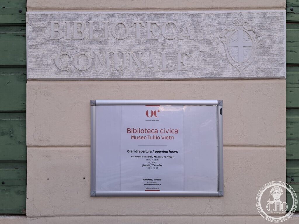 A stone tablet carved Biblioteca Comunale with a coat of arms, above a framed notice with the opening hours of the library and of the Museo Tullio Vietri