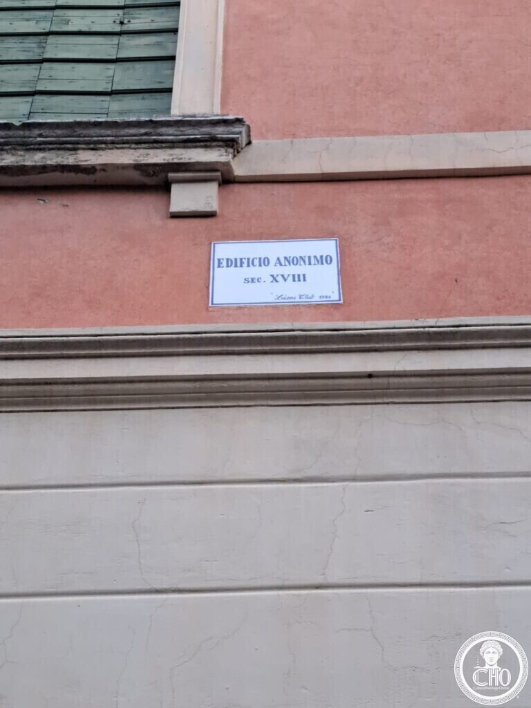 A small marble plaque on an orange wall below a green shutter, reading Edificio anonimo, sec. XVIII, Lions Club