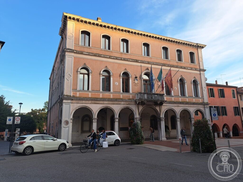 A three-storey orange palace with a ground-floor portico of seven round arches, round-arched windows and a central balcony with three flags, on a pedestrian square