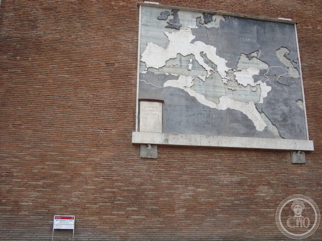 A large grey marble map of the Mediterranean set on a plain brick wall, the Roman Empire picked out in white stone, with a small inscribed panel at the lower left and two stone eagles beneath