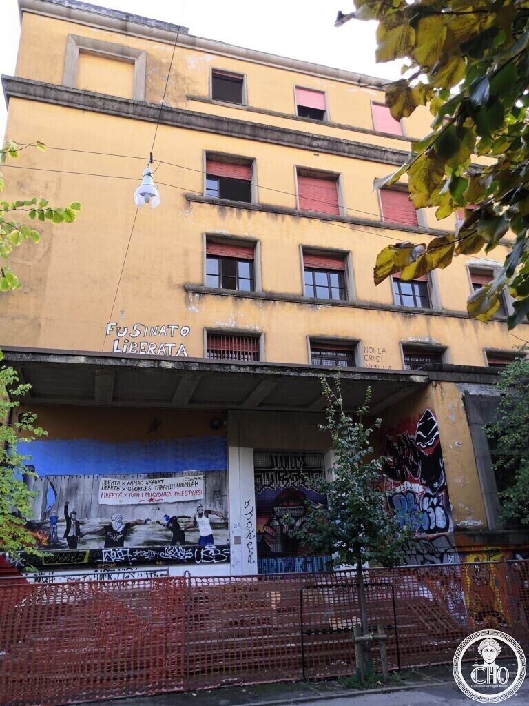 A tall yellow building with rows of windows and red shutters, a concrete canopy over the entrance, the words Fusinato liberata painted on the wall and murals and graffiti below
