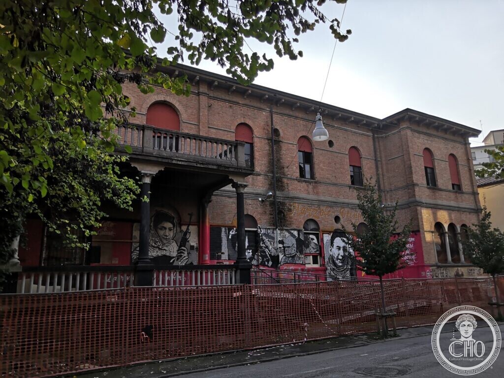 A brick building with round-arched windows and red shutters, a corner loggia on dark columns with a stone balustrade, murals of faces on the walls under it and an orange safety fence along the pavement