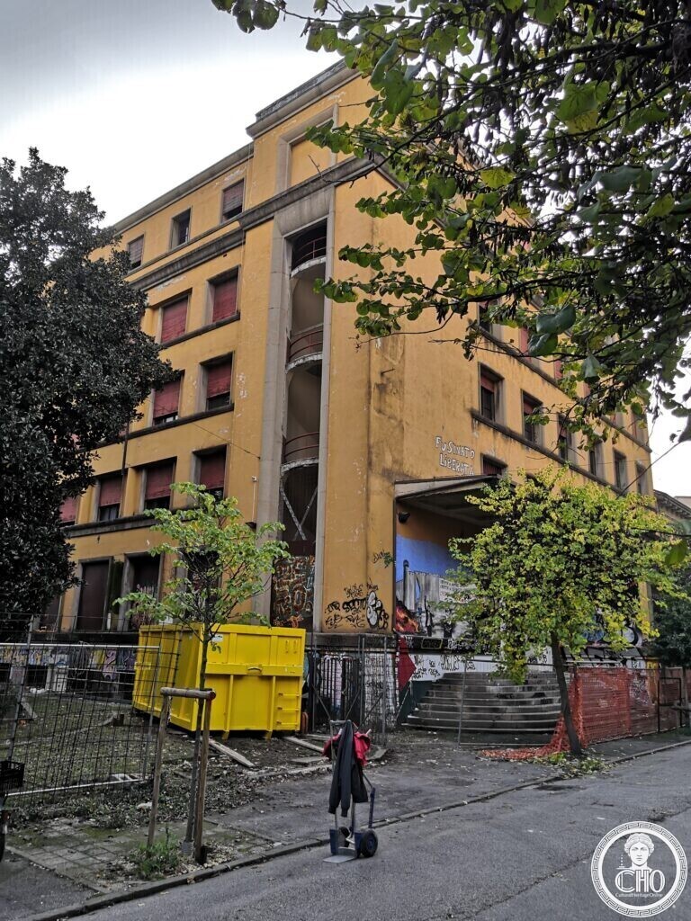 The corner of a tall yellow building with a vertical glazed slot showing a spiral staircase with red railings, a yellow skip and a tree in front