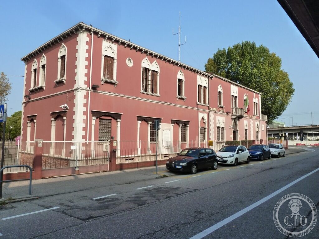 A long two-storey pink building with white stone trim, Venetian Gothic pointed windows, a balcony with the Italian flag and the inscriptions Caserma Corfù and Guardia di Finanza on the front, cars parked along the street and a railway flyover behind