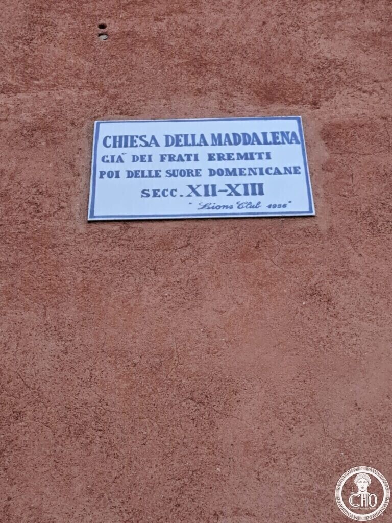 A white plaque with blue lettering on a red wall reading Chiesa della Maddalena, gia dei frati eremiti, poi delle suore domenicane, secc. XII-XIII