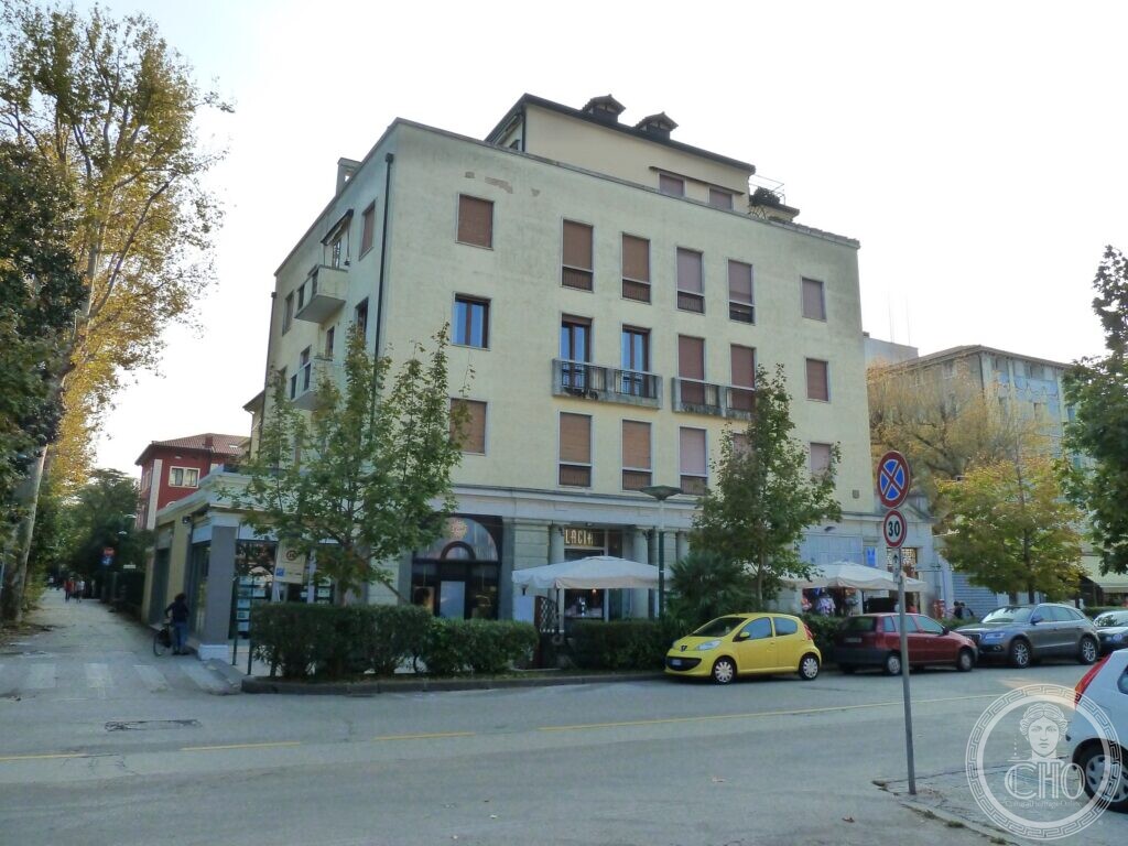 A plain four-storey yellow apartment block with rows of windows and brown shutters, two dormers on the roof, a ground floor of shops with grey stone piers, a restaurant sign and parasols, trees and parked cars on a street corner
