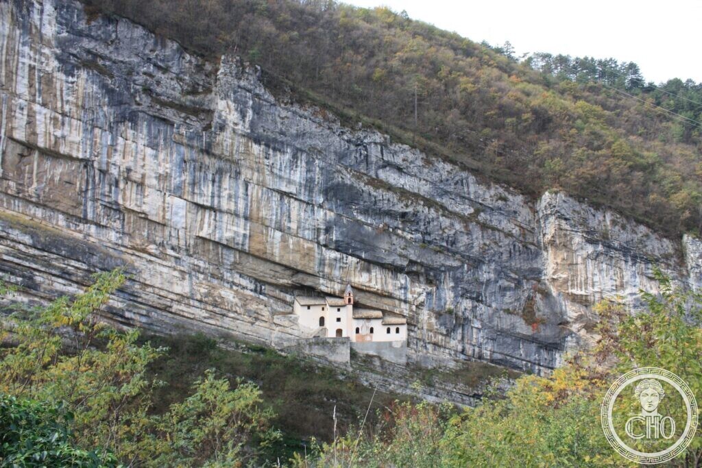 A huge diagonally bedded limestone cliff with a tiny white church complex on a ledge halfway up, autumn woods below and above