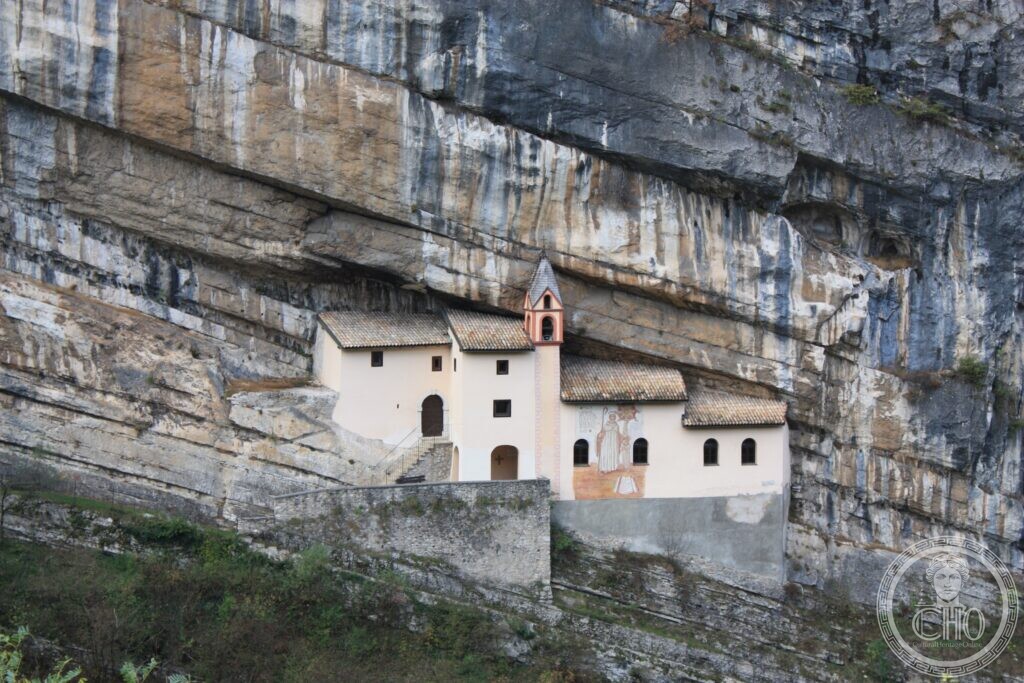 A small white church with a red bell-gable and a painted saint on its wall, built on a ledge under a vast overhanging limestone cliff