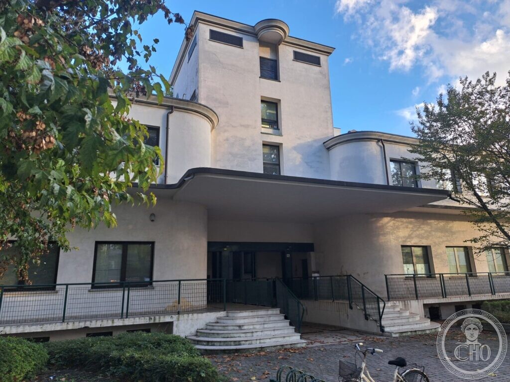 A white modernist building with a flat canopy over a central entrance reached by two curved flights of steps, two rounded wings and a square tower with a rounded corner rising above