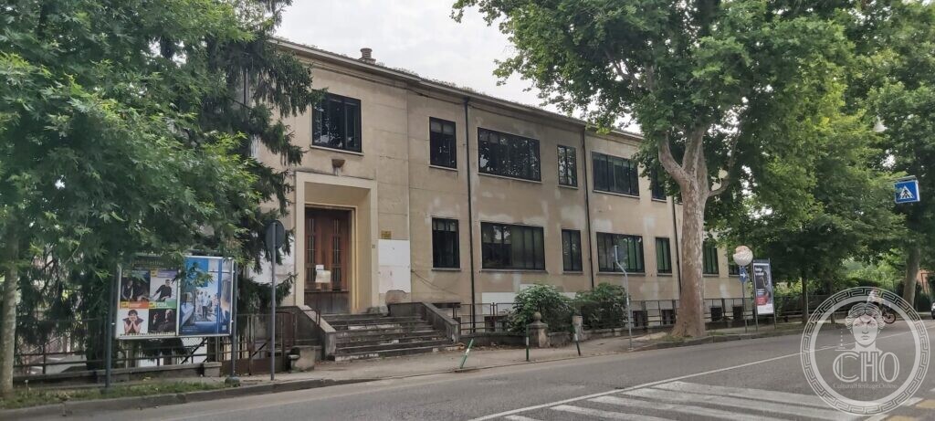 A plain two-storey school building in pale plaster with large rectangular windows, a central doorway framed in stone with a short flight of steps, and plane trees along the road in front