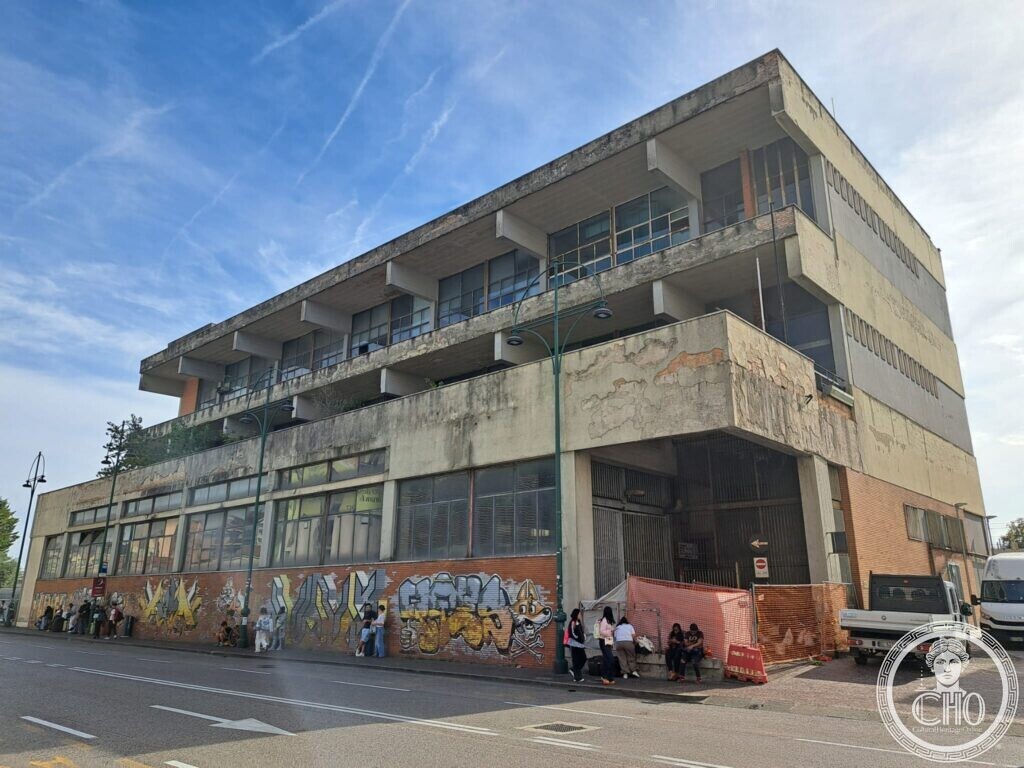 The long concrete building seen from the square with its window bands and balconies, a white van and parked cars in front