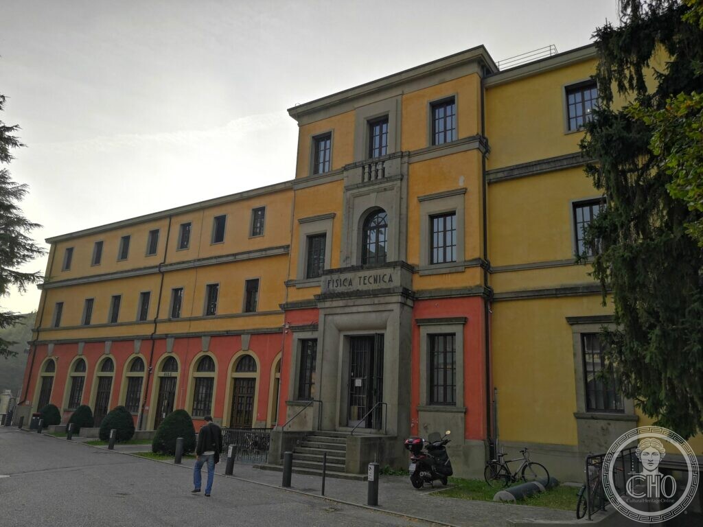 A long yellow university building with a red ground floor of round-arched windows, a taller central block with a stone-framed door, a balustrade above it and the inscription Fisica Tecnica, a man walking past and a scooter parked by the steps