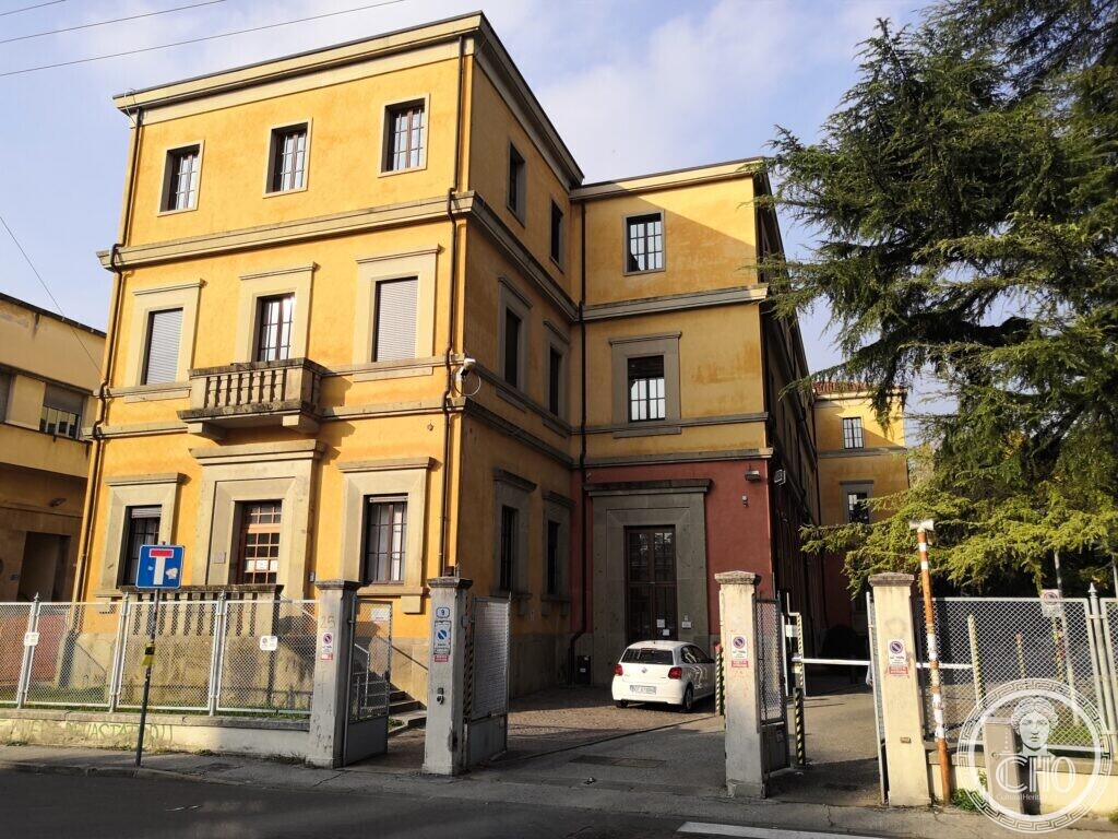 A three-storey yellow building with stone-framed windows, a stone portal with a balcony above it, a lower red wing with a stone-framed door, and an open gate with a parked car