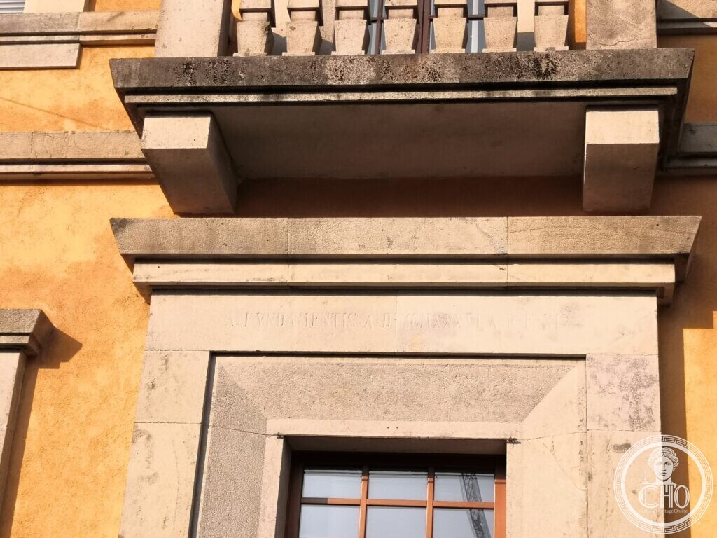 Close-up of a stone lintel under a balcony, with a line of small incised Roman capitals reading A fundamentis A.D. MCMXXXVI A.R.F. XIV