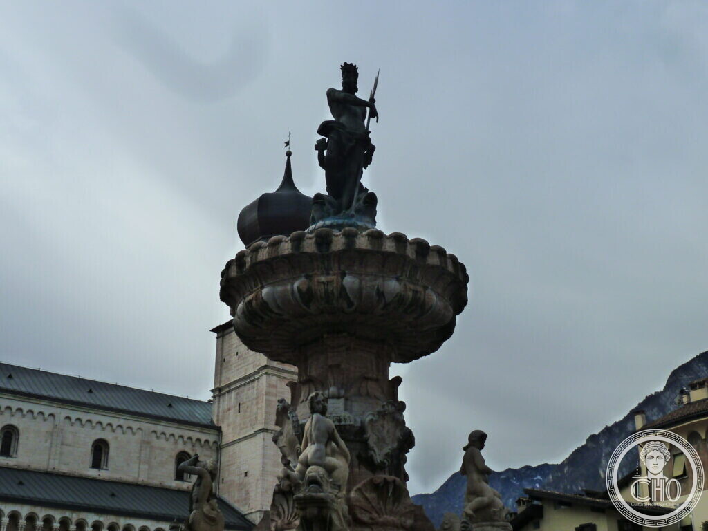 A low-angle close-up of the dark bronze Neptune statue raising a trident atop the fountain, with a church tower and mountains behind.