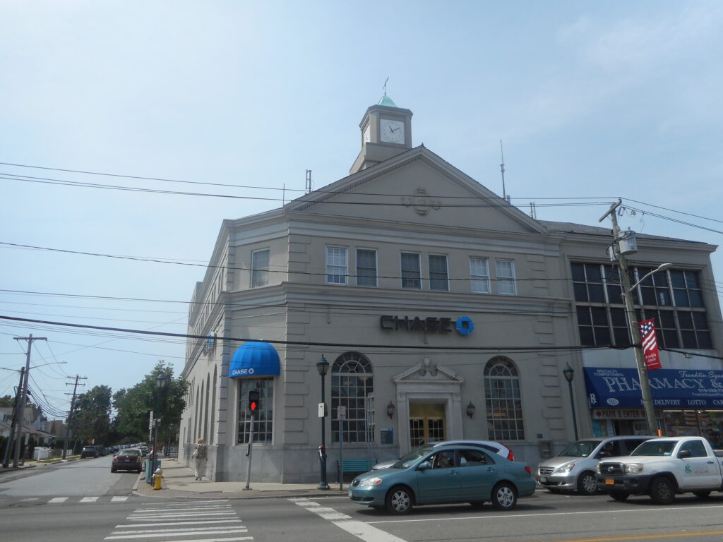 A two-storey bank building in pale stone on a street corner, with a pediment carrying a carved oval cartouche, a white clock lantern with a green dome above it, round-arched windows at ground level and a Chase sign over the entrance