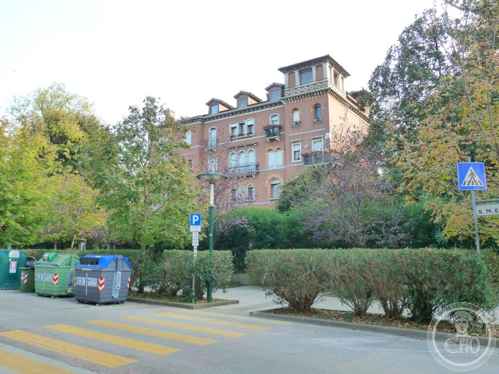 A four-storey apartment building in terracotta plaster with pointed Venetian windows, small iron balconies with flower boxes, a decorated cornice and a row of dormers with a larger central one, half hidden by trees and a hedge, with a street sign reading S. M. E. on the right