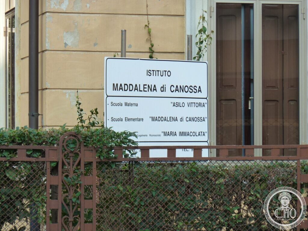 A white board at the gate reading Istituto Maddalena di Canossa, Scuola Materna Asilo Vittoria, Scuola Elementare Maddalena di Canossa, Maria Immacolata, behind an iron railing with ivy