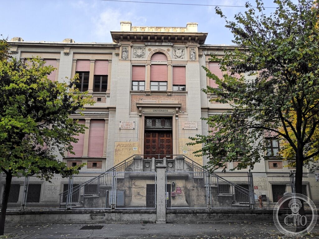 A two-storey institutional building in pale stone and brick with pink shutters, a central bay with a round-arched window, two coats of arms and two statues in niches, the word Università over the door, and a double flight of steps behind a railing