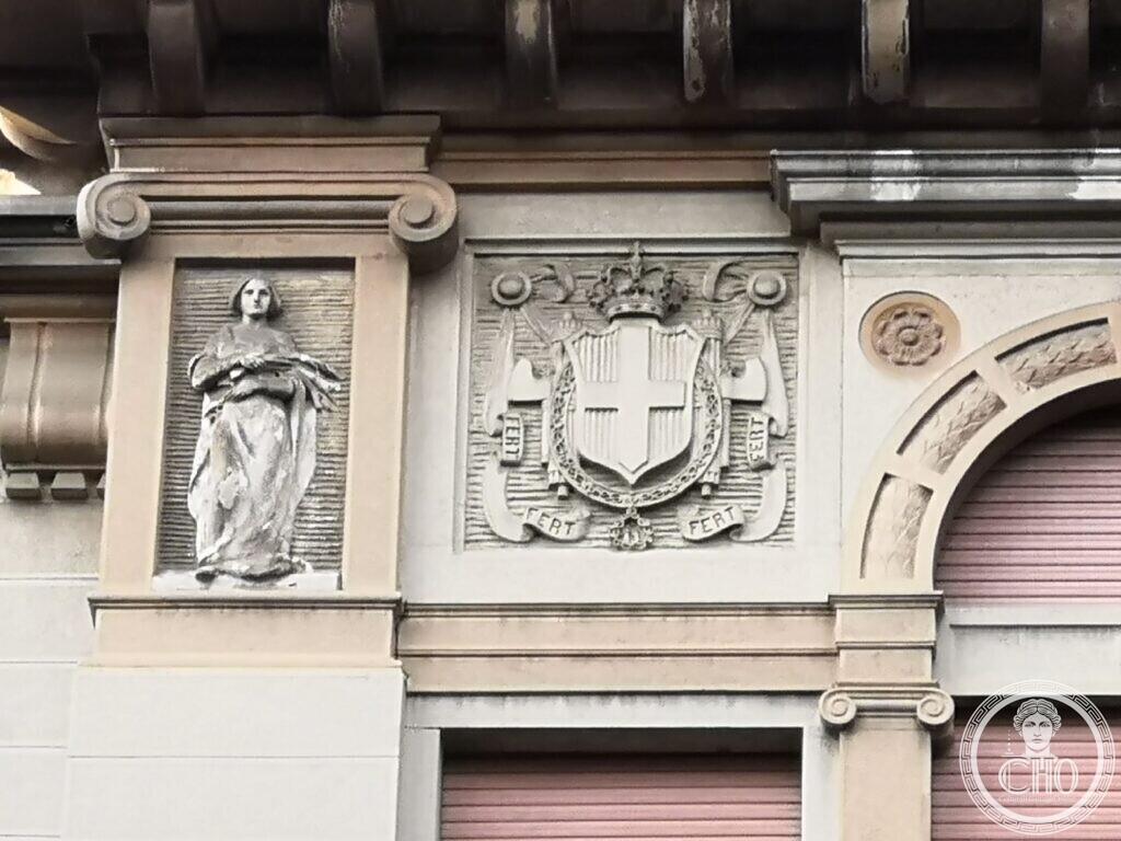 Close-up of a carved stone panel with a crowned shield bearing a cross inside a collar, ribbons lettered FERT, and a draped standing figure in a niche to the left under a stone cornice
