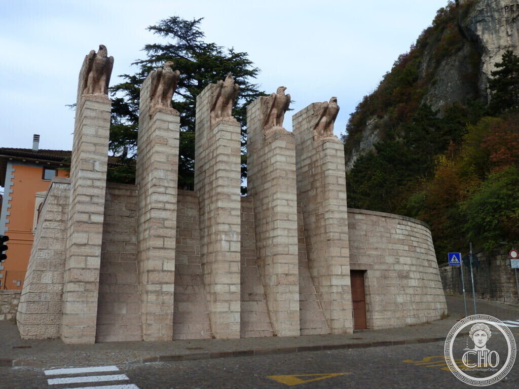 The full monument with curved wall and five eagles, seen from the side near a rocky hillside.
