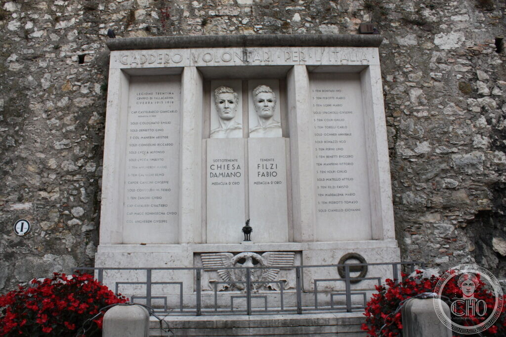 A white marble memorial set against a rough stone wall, with two carved male busts side by side under an inscribed architrave and long lists of names on either side