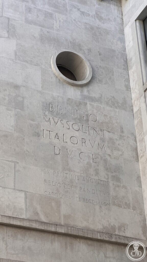 A wall with a round window and carved Latin inscriptions honoring Mussolini