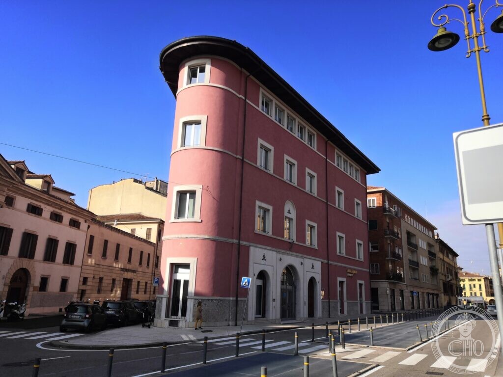 A four-storey pink building shaped like a wedge, with a rounded prow of stacked windows at the corner, white window frames, a grey stone plinth and three arched doorways, on a street corner under a deep blue sky