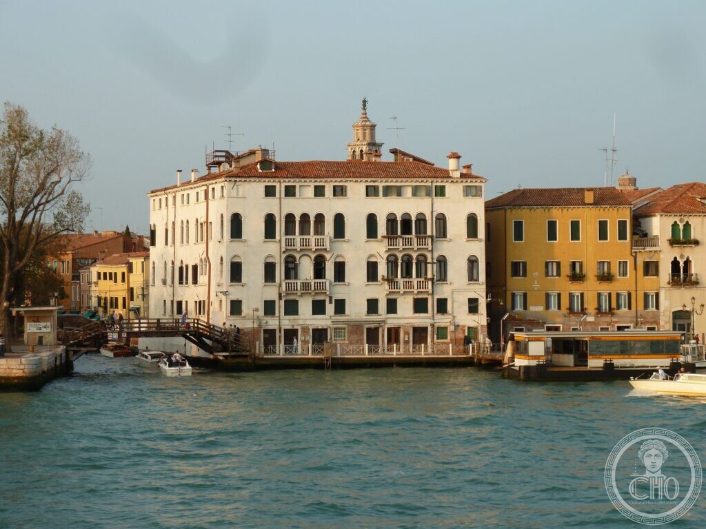 A white four-storey palace with Gothic pointed windows and small balconies on the Giudecca Canal, a wooden bridge on the left, a vaporetto landing stage on the right and a bell tower rising behind