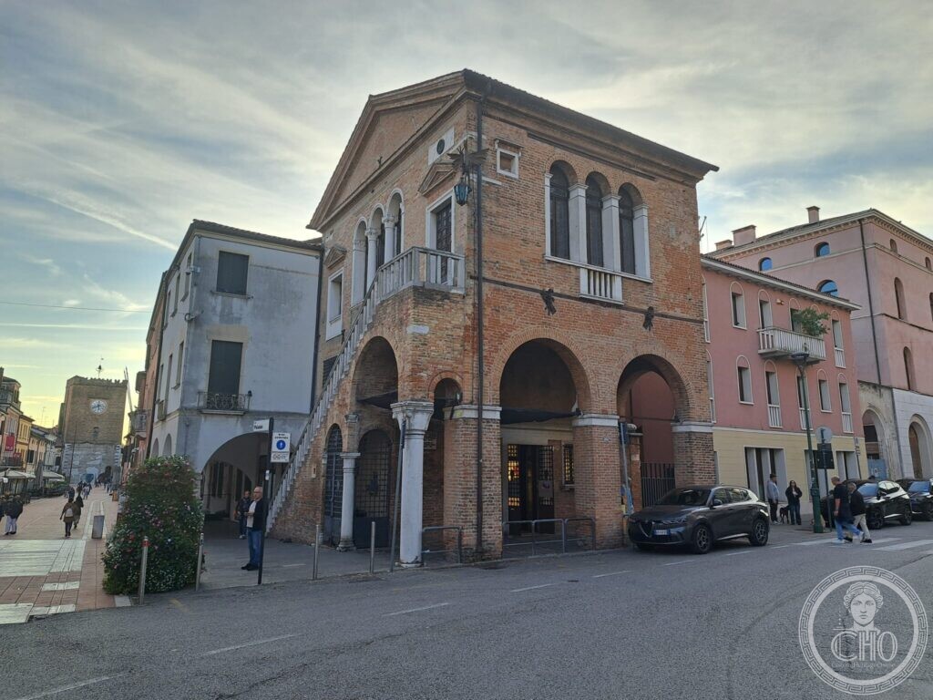 A brick two-storey building with a triangular pediment, a three-light arched window above two round arches, and an external stone staircase, at the end of a pedestrian street with a clock tower in the distance