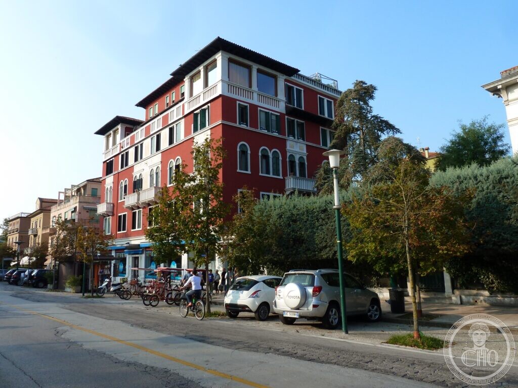 A tall red apartment building with white stone trim, round-arched windows, small balconies with flower boxes, an open loggia under the eaves at the top with a roof terrace, and shops with blue awnings at street level, seen from across a tree-lined street with parked cars and bicycles