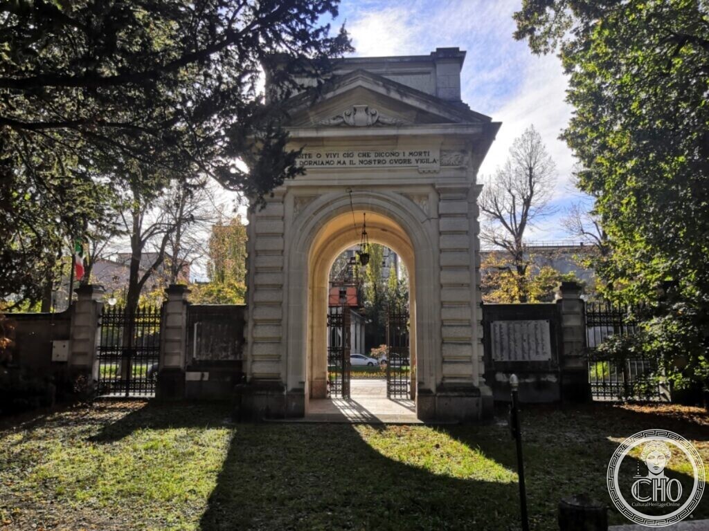 The same stone gateway seen from inside the park, with an inscription on the frieze, the iron gate open and the road and trees beyond