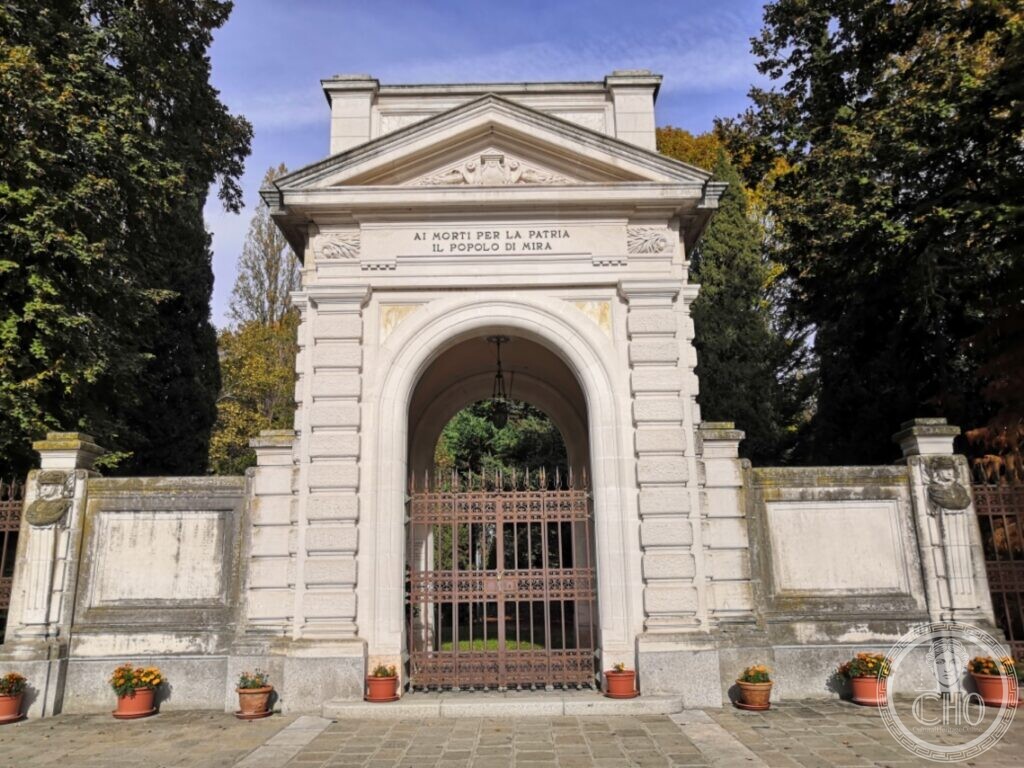 A white stone gateway with a triangular pediment and a round arch closed by an iron gate, the inscription Ai morti per la patria il popolo di Mira on the frieze, flanked by low walls with blank plaques, flower pots on the pavement and tall trees behind