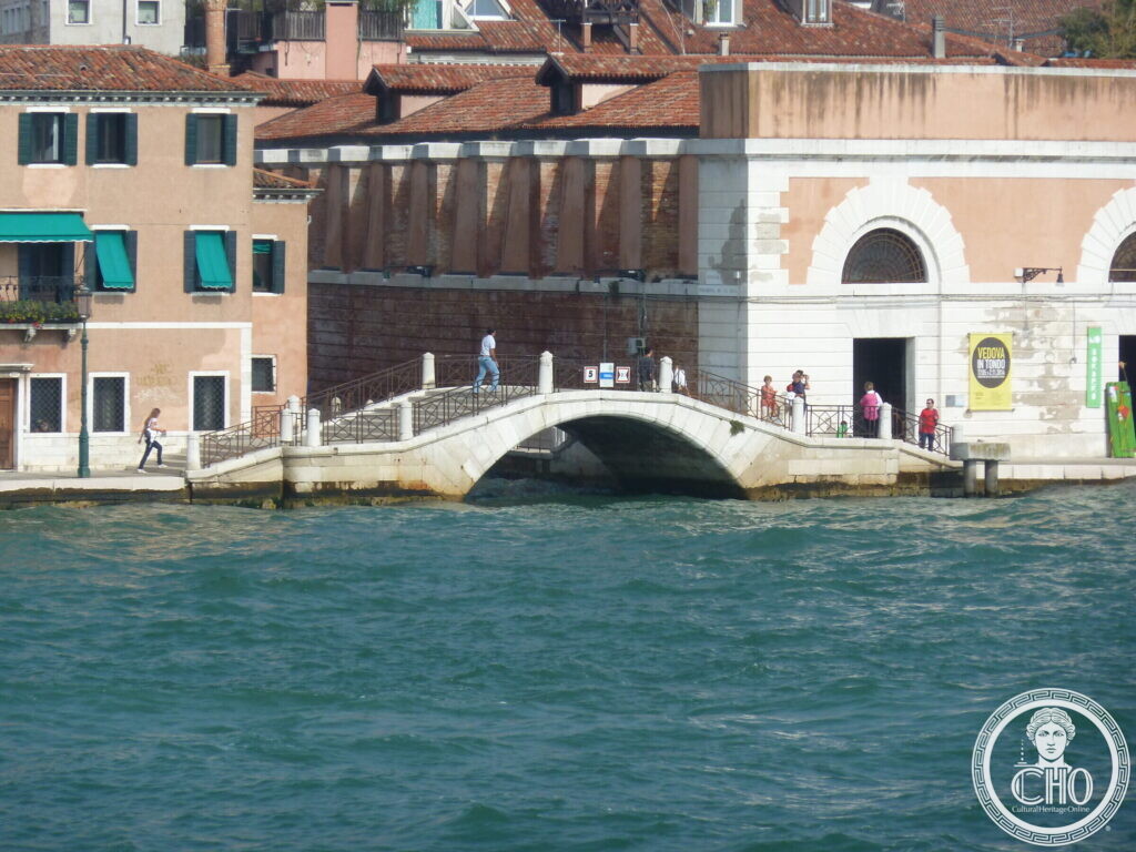 A small stone arch bridge with brick parapets crossing a narrow canal beside the long brick front of the salt warehouses on the Zattere