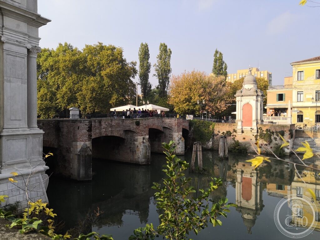 A brick bridge of three arches with stone cutwaters over a still canal, people under white umbrellas on the parapet, a small Baroque shrine with a dome on the far bank, poplars and plane trees behind, and the stone pier of a gate in the foreground