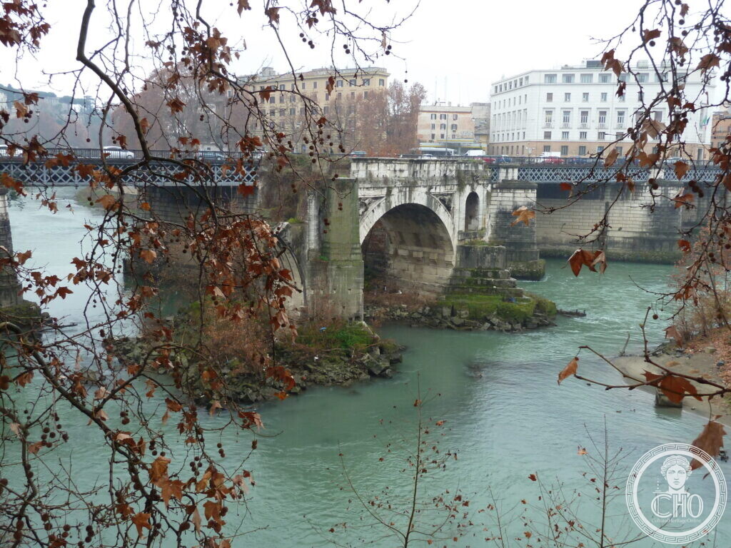 The lone arch of the Ponte Rotto in the river with a modern iron bridge and tall houses behind, autumn leaves in the foreground