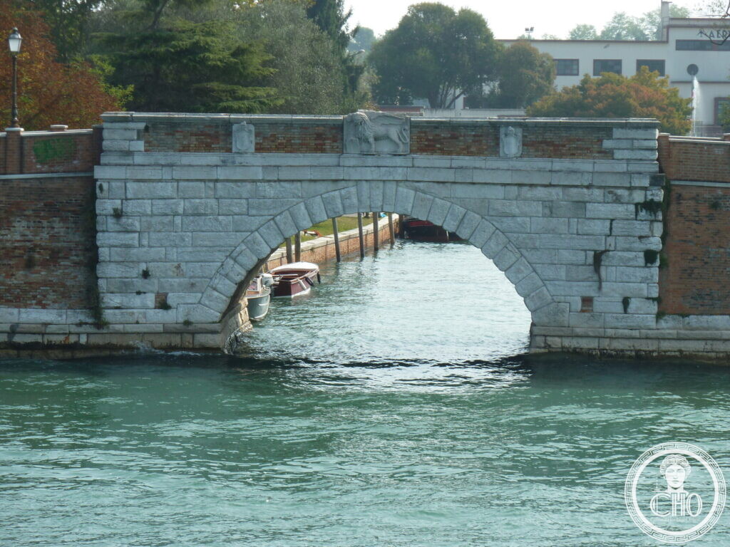 A single-arched stone bridge over a canal with a brick parapet, a carved winged lion and two shields in the middle, boats moored under the arch