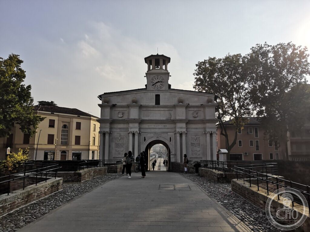 A white stone city gate with paired columns, a round archway, two coats of arms, the inscription Omnium Sanctorum on the frieze and a small clock tower with a lantern on top, at the end of a cobbled bridge with low brick parapets