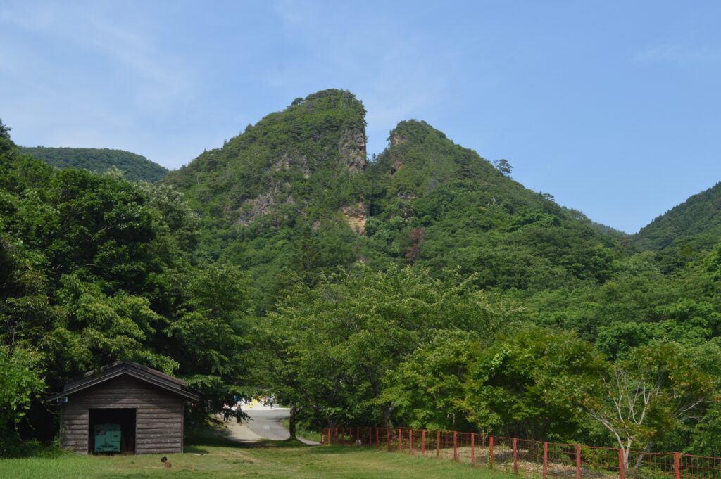 The wooded ridge above the Aikawa mine on Sado, with the notch of the Doyu-no-Warito cut in the rock face and a small timber mine building on the grass below