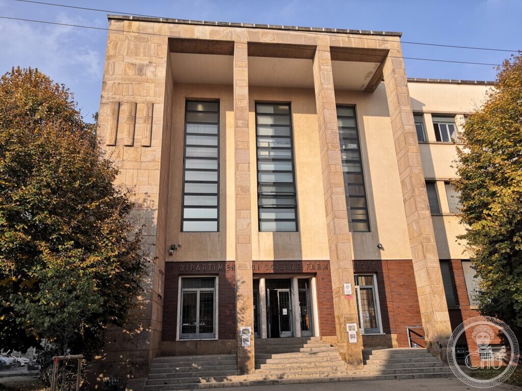 A tall stone-faced building front with three giant piers framing two full-height strips of windows, a dark brick base with the inscription Dipartimento di Scienze Farmaceutiche, a flight of steps to the glazed entrance, and autumn trees on either side