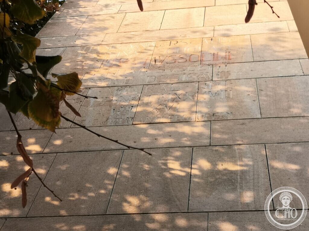 Lines of worn Roman capitals carved into pale stone blocks, one line in red, seen through the leaves of a lime tree in dappled light